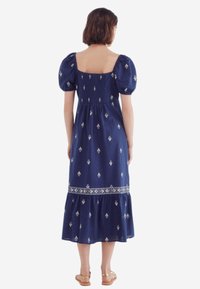 Robe en coton bleu marine avec manches bouffantes, encolure carrée et jupe à volants. Présente une broderie blanche et un dos froncé.