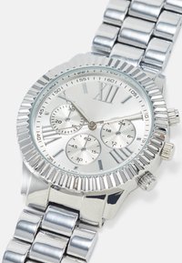 Silversmycke med en strukturerad bezel, romerska siffror som markörer, tre subdials och ett armband i rostfritt stål. Glansig yta genomgående.