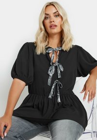 Blusa nera con maniche a sbuffo, cintura in vita e design a chiave, accentuata da un cravattino a contrasto con motivo. Indossata con jeans grigi chiari.