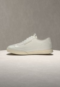 Άσπρο low-top αθλητικό παπούτσι με κορδόνια μπροστά, ανοιχτό μπεζ σόλα και λογότυπο "EMPORIO ARMANI" στη πλευρά, εμφανίζεται σε ουδέτερη επιφάνεια.