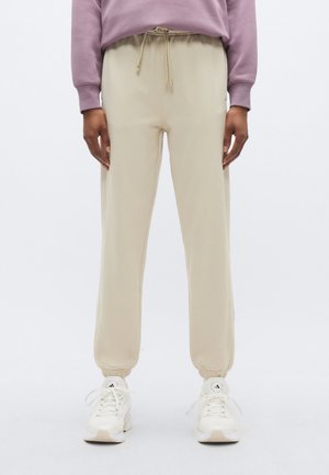 Beige joggebukser laget av mykt materiale, med snøring i midjen, elastiske mansjetter og sidelommer, kombinert med hvite joggesko.