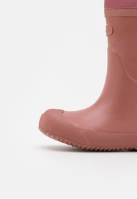 Roze rubberen laars met een gestructureerde zool, ronde neus en een glad bovenwerk. Heeft een contrasterende stofrand aan de bovenkant.