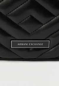 Μαύρη δερμάτινη επένδυση με διαγώνια ραφή και ορθογώνια μαύρη μεταλλική πλάκα με χαραγμένη την επιγραφή "ARMANI EXCHANGE" σε λευκό χρώμα.