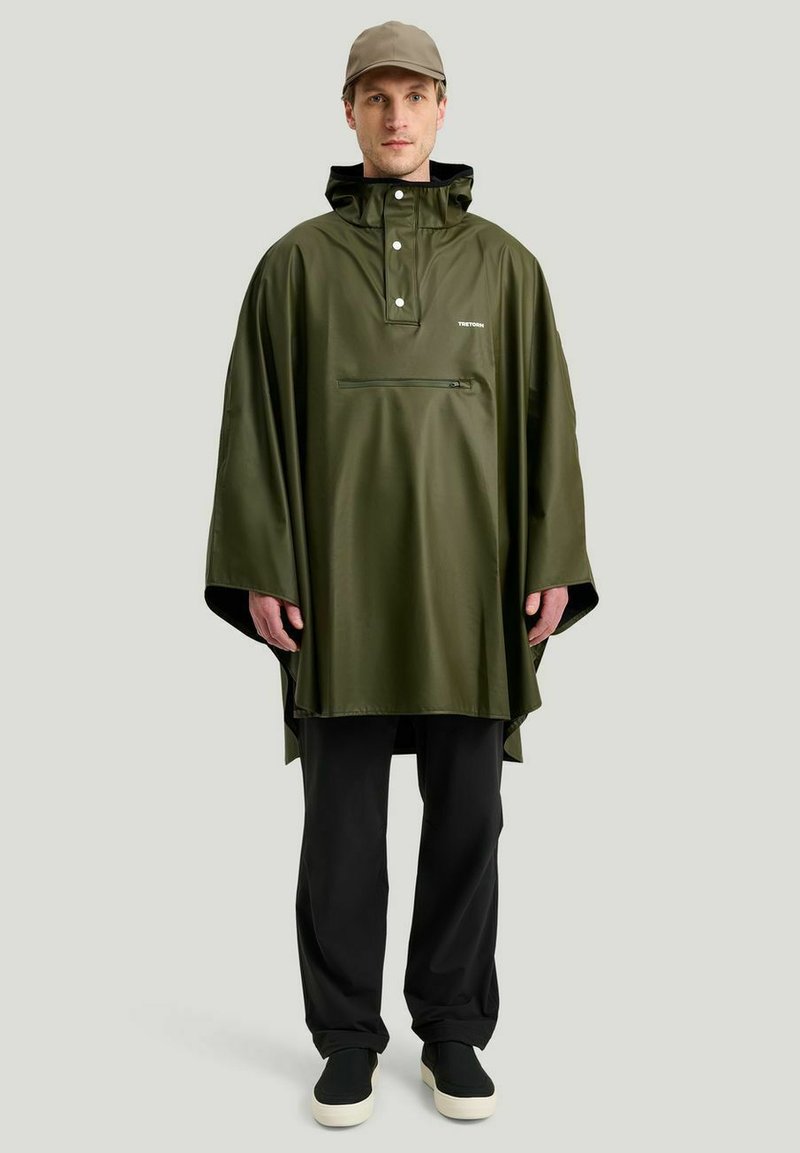 Vihreä vedenpitävä poncho, suuri huppu, etutasku vetoketjulla, kiiltävä pinta, käytetty mustien housujen ja liukuvaisten kenkien kanssa, rento tyyli.