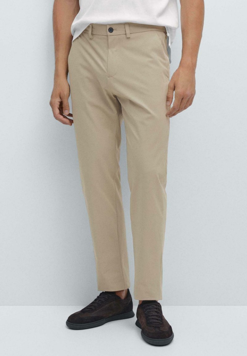 Pantalons beige slim-fit portés avec une chemise blanche et des baskets en daim noires, coupés à la cheville devant un fond clair.