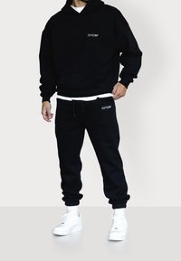Hombre que lleva una sudadera negra con capucha y pantalones deportivos a juego con logotipos blancos, zapatillas blancas y calcetines blancos, de pie frente a un fondo liso.