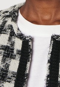 Gros plan sur une personne portant une chemise blanche et une veste à motif noir et blanc texturé avec des bords effilochés.