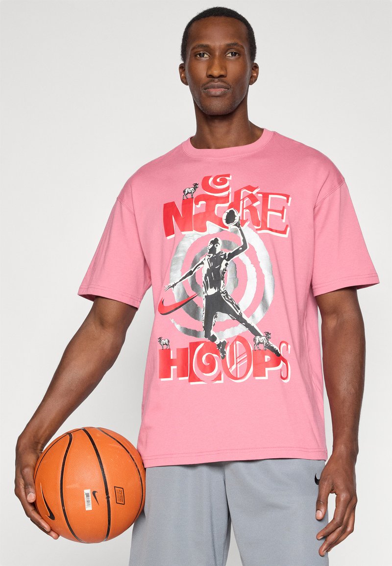 T-shirt di cotone rosa con stampa grafica che presenta un giocatore di basket, il testo "NIKE" e "HOOPS". Modello che tiene un pallone da basket arancione, indossando pantaloni corti grigi.
