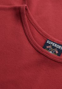 Rød bomulds T-shirt med ribbet rund halsudskæring og en mærketiket med tekst og grafisk design i blå, hvid og gul accent.
