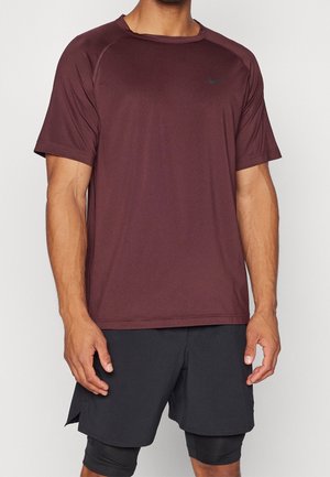 Homme portant un t-shirt de sport bordeaux à manches courtes et un short noir superposé sur un fond clair uni.