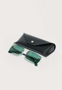 Gafas de sol rectangulares con tinte verde y montura de oro colocadas junto a un estuche cerrado de cuero negro sobre una superficie blanca.