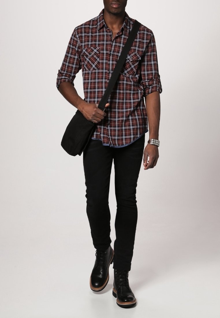 Homme marchant portant une chemise à carreaux rouge, un pantalon noir slim, des bottes en cuir noires, un bracelet en argent et portant un sac bandoulière noir.