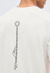 Άτομο που φοράει λευκό πουκάμισο με έναν κάθετο μαύρο σχεδιασμό που απεικονίζει έναν ήλιο, ένα βουνό, και κείμενο που γράφει "ΣΤΗ ΣΙΩΠΗ ΤΗΣ ΦΥΣΗΣ ΒΡΙΣΚΕΙΣ ΤΗΝ ΠΙΟ ΔΥΝΑΤΗ ΠΕΡΙΠΕΤΕΙΑ."