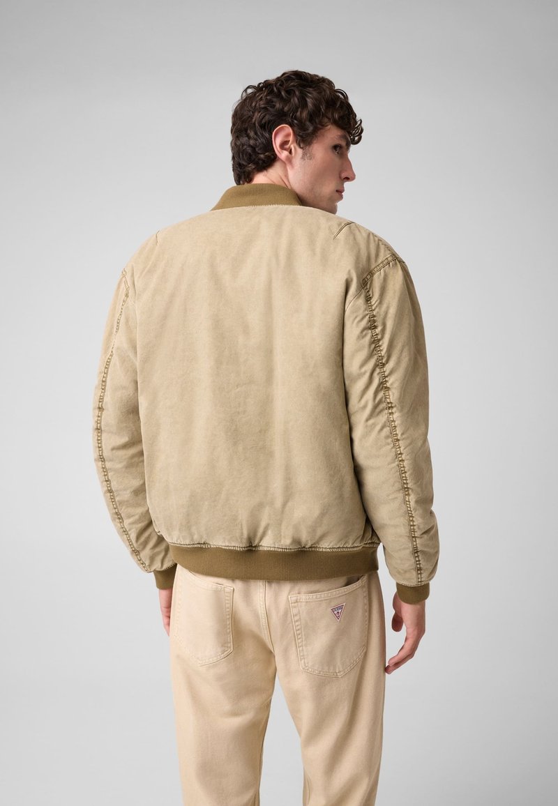 Homme aux cheveux courts et bouclés portant une veste bomber beige et un pantalon beige clair, de dos sur un fond gris clair uni.