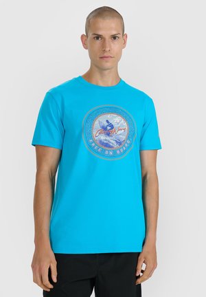 Mężczyzna w jasnoniebieskim T-shircie z grafiką surfingu i napisem "Chasing Waves Free on Board" na jednolitym tle.