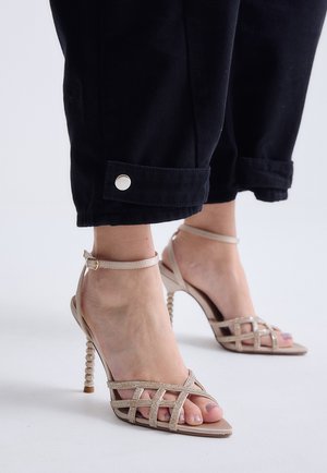 Pies que llevan sandalias nude de tacón alto con tiras y correas en los tobillos, junto con pantalones negros remangados en los tobillos con detalle de botón plateado.