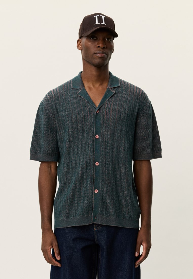 Πλεκτό μπλουζάκι με κοντά μανίκια σε τιρκουάζ απόχρωση, με υφή, ροζ κουμπιά και γιακά. Φοριέται με σκούρο τζιν παντελόνι και καφέ καπέλο.