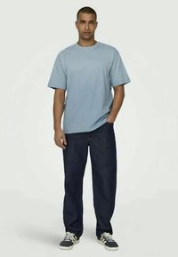 Light blue short-sleeve t-shirt with a relaxed fit, paired with dark denim jeans. The model stands with one hand in his pocket, wearing sneakers.