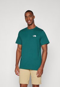 Tricou de bumbac teal cu mâneci scurte și gât rotund, având un logo alb pe partea stângă a pieptului. Croială lejeră, textură netedă.