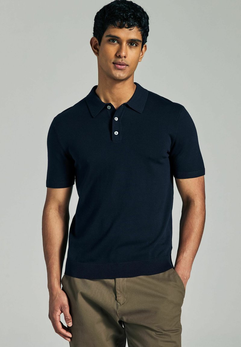 Marineblaue Kurzarm-Poloshirt mit Kragen, Knopfverschluss, gerippten Bündchen und Saum. Weiche, gestrickte Textur, kombiniert mit khakifarbenen Hosen.