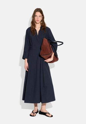 Młoda kobieta w długiej granatowej sukience i czarnych sandałach, trzyma dużą brązową zamszową torebkę na białym tle.