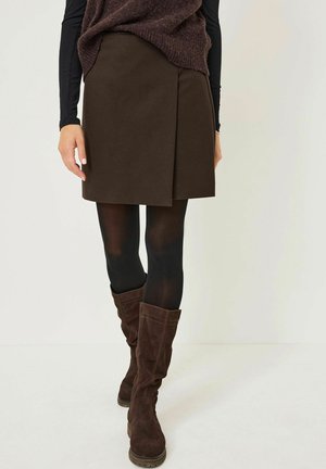 Bruine stoffen rok met een wikkelontwerp, gecombineerd met zwarte panty’s en bruin suède laarzen tot de knie. De outfit heeft een gestructureerd donker trui.