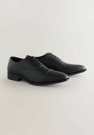 Chaussures habillées en cuir noir avec une finition texturée, un design à bout caps, et une semelle en caoutchouc plate, dotées d'un laçage et d'un col rembourré.