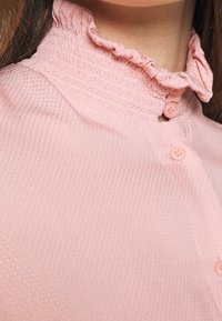 Pink shirt with a textured fabric, featuring a button-up front and a ruffled collar. Five matching buttons accent the neckline.