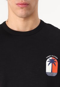 Juodas marškinėliai su apvaliu iškirpimu, su apvalia etikete, kurioje pavaizduota palmės dizainas oranžine, mėlyna ir balta spalvomis, ir tekstas "TOMMY HILFIGER".