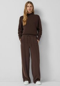 Brązowy sweter z wysokim kołnierzem, w połączeniu z luźnymi brązowymi spodniami z troczkiem w pasie. Całość dopełniają białe trampki.