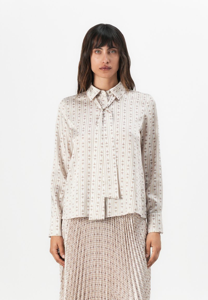 Blouse à manches longues en tissu léger ; présente un col, un détail à nouer et un tissu beige avec des motifs géométriques verticaux.