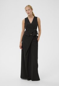 Čierna bezrukávová zavinovacia blúzka s výstrihom do V, zaisťovaná bočnou prackou, doplnená o široké nohavice so zodpovedajúcim vzorom a záhybmi.