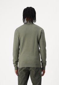 Olivgrüner Pullover mit steilem Kragen, gerippten Bündchen und glatter Strickstruktur. Getragen über passenden grünen Cargo-Hosen.