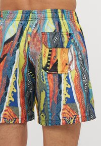 Shorts de bain colorés avec des motifs abstraits en bleu, jaune, orange et noir. Comprend une poche de accent à l'arrière.