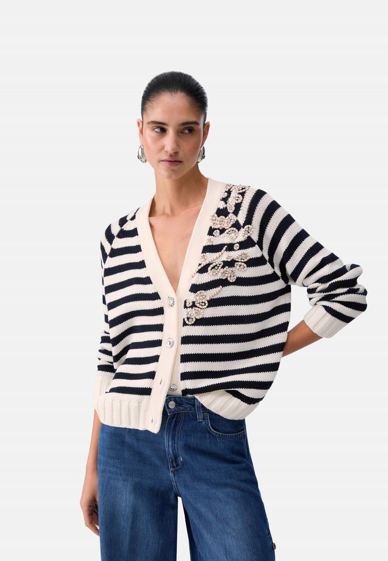 Vrouw met naar achter gestyled haar, gekleed in een zwart-wit gestreepte cardigan met bloemrijke borduursels en een blauwe spijkerbroek, staand met één hand op de heup.