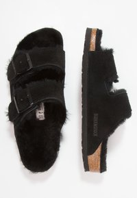 Pantofole nere in suede con interno in pelliccia sintetica, due cinturini regolabili con fibbie nere e una soletta in sughero. La suola presenta una presa testurizzata.