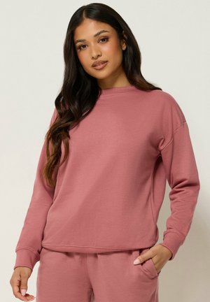Frau mit langen dunklen Haaren, die einen mauvefarbenen Sweatshirt mit langen Ärmeln und passende Hosen trägt, steht mit einer Hand in der Tasche.
