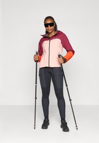 Veste multicolore avec des sections bordeaux, rose et orange, leggings noirs avec poches et bottes de randonnée noires ; tenant des bâtons de randonnée.