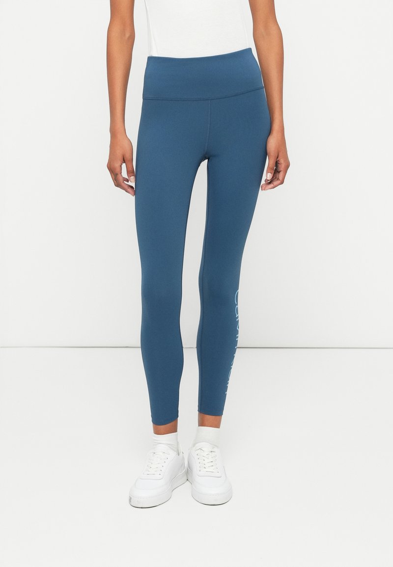 Leggings justos, de cintura alta, em um tecido azul profundo, com uma textura suave e um detalhe de logótipo branco na parte inferior da perna.