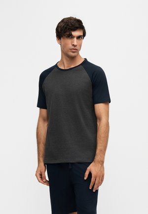 Homme aux cheveux courts et foncés portant un t-shirt à manches raglan gris foncé et noir et un short noir, debout devant un fond clair uni.