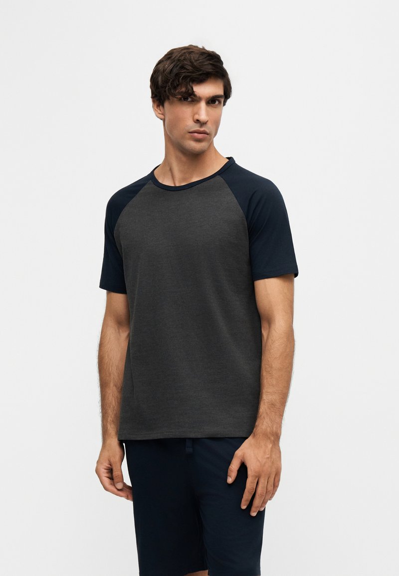 Homme aux cheveux courts et foncés portant un t-shirt à manches raglan gris foncé et noir et un short noir, debout devant un fond clair uni.