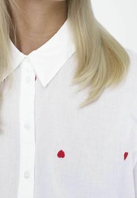 Chemise à col blanc en tissu léger, ornée de cœurs rouges brodés. Les boutons sont transparents et ronds, avec une texture lisse.