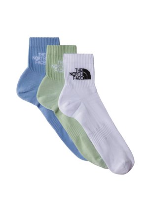 Młody mężczyzna w granatowej koszulce polo, beżowych spodniach cargo, zielonych sneakersach i czapce w kolorze czarnym, trzymający białą torbę tote, stojący na jednolitym tle.