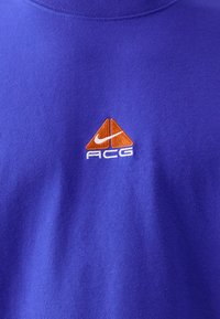 Lila pamut póló, hímzett narancssárga és fehér logóval, amelyen két háromszög és "ACG" felirat található, kerek nyakkal és puha tapintással.