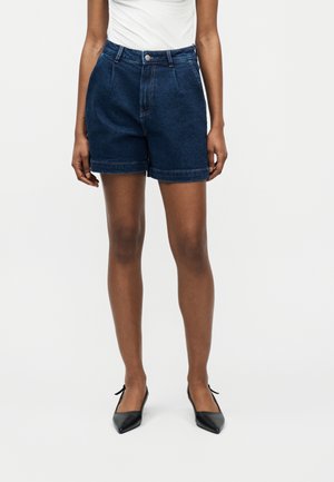 Γυναίκα που φοράει πλισέ σκούρο μπλε τζιν σορτς, λευκό τοπ και μαύρα μυτερά τακούνια, στέκεται μπροστά σε απλό ανοιχτό φόντο.