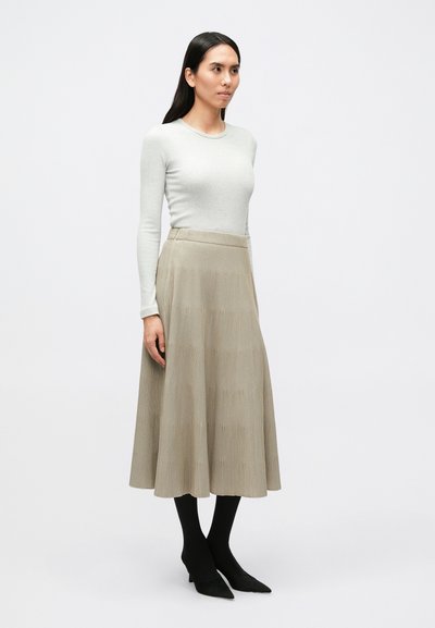 Ανοιχτό γκρι μπλούζα με μακριά μανίκια συνδυασμένη με μπεζ πλισέ φούστα μεσαίου μήκους. Μαύρες καλσόν και μαύρα τακούνια με μυτερή μύτη ολοκληρώνουν την εμφάνιση.