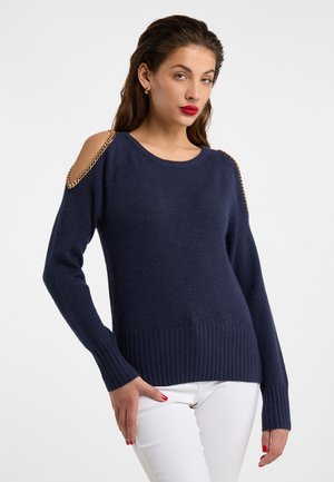 Frau trägt einen marineblauen Pullover mit goldenen Kettendetails an ausgeschnittenen Schultern und weiße Hose, steht vor einfarbig hellem Hintergrund.