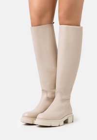 Bottes en cuir beige jusqu'aux genoux avec de lourds semelles en caoutchouc blanc. Texture lisse avec des détails de coutures subtils et un design à bout rond.