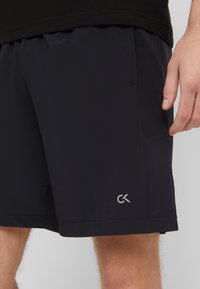 Svarta sportshorts i lätt tyg med elastisk midja, med en CK-logotyp på nedre vänstra delen, slät yta och utan synliga mönster.