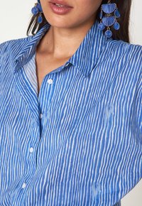 Camicia a bottoni a righe blu con linee verticali e colletto, realizzata in un tessuto leggero e texturizzato. Bottoni bianchi e vestibilità comoda.